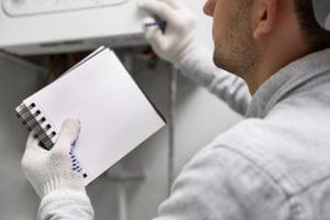 Heating Repair Person in gloves with a notebook, inspecting a boiler or appliance with a screwdriver.