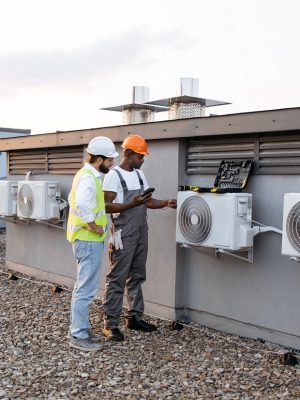 Group of multicultural construction workers on roof inspecting cooling system together using testing tool. Large and gray air conditioning unit with multiple ventilation openings and pipes.
